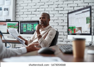 Two Diverse Colleagues Traders Talking To Each Other While Sitting In The Office In Front Of Multiple Computer Screens. Stock Trading, People, Business Concept. Selective Focus