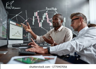 Two Diverse Colleagues Traders Talking To Each Other, Looking At Graphs While Sitting In The Office In Front Of Multiple Computer Screens. Stock Trading, People, Business Concept.