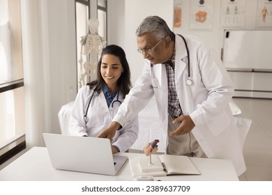 Two diverse doctors discussing online medical application, using laptop at workplace together. Indian elderly medical professional teacher, mentor training young Latin intern in clinic office - Powered by Shutterstock