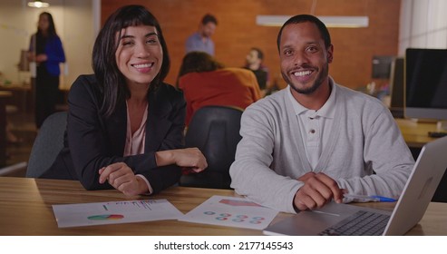 Two Diverse Employees At Work. An African Man Talking With A Hispanic Female Brazilian Colleague In Front Of Laptop Computer