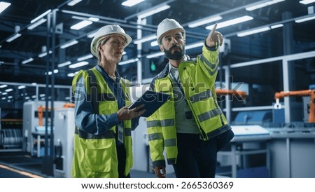 Two Diverse Engineers Walking, Using Tablet Computer at an Industrial Machinery Factory. Caucasian Female and Hispanic Male Specialists Chatting and Monitoring Robots on a Modern Assembly Line.