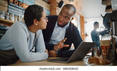 Two Diverse Entrepreneurs Have A Team Meeting In Their Stylish Coffee Shop. Barista And Cafe Owner Discuss Work Schedule And Menu On Laptop Computer. Multiethnic Female And Male Restaurant Employees.