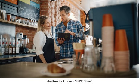 Two Diverse Entrepreneurs Have A Team Meeting In Their Stylish Coffee Shop. Barista And Cafe Owner Discuss Work Schedule And Menu On Tablet Computer. Young Female And Male Restaurant Employees.