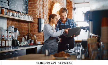 Two Diverse Entrepreneurs Have A Team Meeting In Their Stylish Coffee Shop. Barista And Cafe Owner Discuss Work Schedule And Menu On Laptop Computer. Multiethnic Female And Male Restaurant Employees.