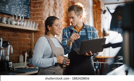 Two Diverse Entrepreneurs Have A Team Meeting In Their Stylish Coffee Shop. Barista And Cafe Owner Discuss Work Schedule And Menu On Laptop Computer. Multiethnic Female And Male Restaurant Employees.