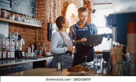 Two Diverse Entrepreneurs Have A Team Meeting In Their Stylish Coffee Shop. Barista And Cafe Owner Discuss Work Schedule And Menu On Laptop Computer. Multiethnic Female And Male Restaurant Employees.