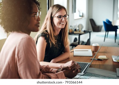 Two diverse female designers smiling while working together at a boardroom table during a meeting in a modern office