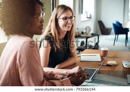 Two diverse female designers smiling while working together at a boardroom table during a meeting in a modern office