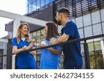 Two diverse female and one male healthcare workers in scrubs take a coffee break outside a medical facility, engaging in a lively conversation.