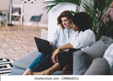 Two Diverse Friends Relaxing On Comfortable Couch And Watching Film Online On Laptop Device Using Home Wireless Internet.Multicultural Young Women Working Together On Common Blog At Computer