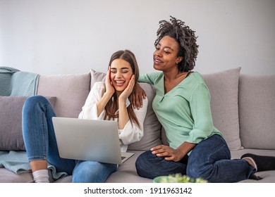 Two Diverse Friends Relaxing On Comfortable Couch And Watching Film Online On Laptop Device Using Home Wireless Internet.Multicultural Young Women Working Together On Common Blog At Computer