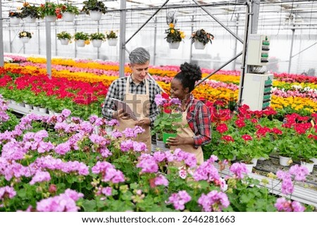 Two diverse greenhouse workers discussing a potted plant, surrounded by vibrant flowers of various colors.