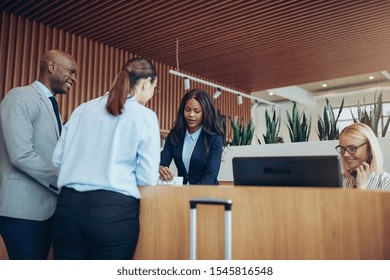 Two Diverse Guests Smiling And Talking To A Concierge While Checking In Together At The Reception Counter Of A Hotel