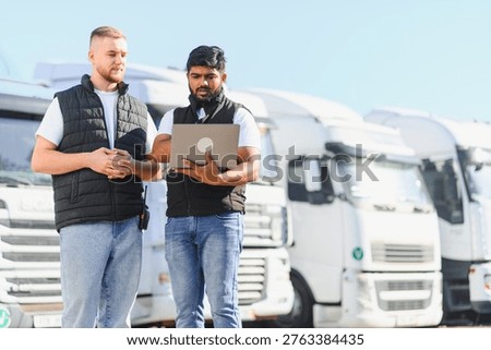 Two diverse logistics managers discussing shipment plans, using a laptop outdoors near a fleet of semi trucks