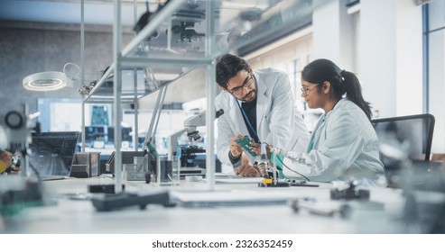 Two Diverse Multiethnic Colleagues Chatting in Laboratory, Working on a Circuit Board for a Computer. Young Indian Scholar Talking with a Scientist About Inserting Different Microchips and Conductors - Powered by Shutterstock