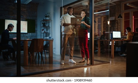 Two Diverse Multiethnic Colleagues Have A Conversation In A Meeting Room Behind Glass Walls In An Agency. Female Creative Director And African American Project Manager Discuss Work On Laptop Computer.