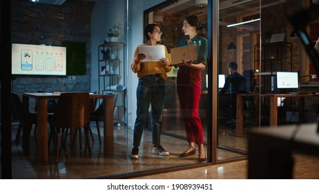 Two Diverse Multiethnic Female Have A Discussion In Meeting Room Behind Glass Walls In An Agency. Creative Director And Project Manager Compare Business Results On Laptop And App Designs In An Office.