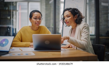 Two Diverse Multiethnic Female Have A Discussion In Meeting Room Behind Glass Walls In An Agency. Creative Director And Project Manager Compare Business Results On Laptop And App Designs In An Office.