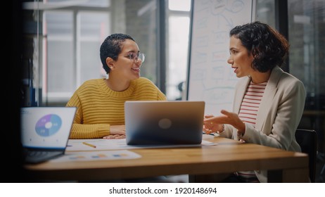 Two Diverse Multiethnic Female Have A Discussion In Meeting Room Behind Glass Walls In An Agency. Creative Director And Project Manager Compare Business Results On Laptop And App Designs In An Office.