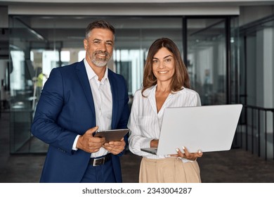 Two diverse partners, colleagues, team of confident professional business people work together. Mature Latin business man and European business woman working on project using digital gadget in office. - Powered by Shutterstock