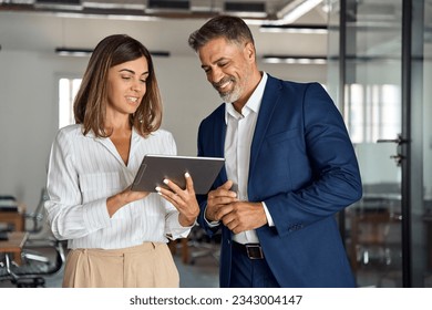 Two diverse partners walking mature Latin business man and European business woman discussing project on tablet in office. Two colleagues of professional business people working together at workspace. - Shutterstock ID 2343004147