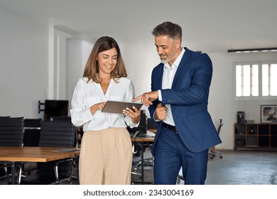 Two diverse partners walking mature Latin business man and European business woman discussing project on tablet in office. Two colleagues of professional business people working together at workspace. - Powered by Shutterstock