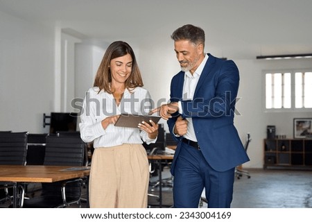 Two diverse partners walking mature Latin business man and European business woman discussing project on tablet in office. Two colleagues of professional business people working together at workspace.