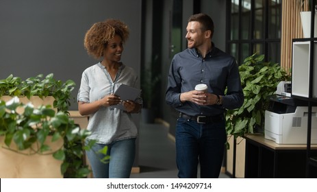 Two diverse professional colleagues talking walking in modern office, happy friendly african female and caucasian male coworkers having conversation discuss project going along business work space