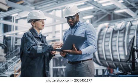 Two Diverse Professional Heavy Industry Engineers Wearing Safety Uniform And Hard Hats Working On Laptop Computer. African American Technician And Female Worker Talking On A Meeting In A Factory.