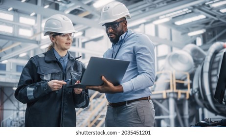 Two Diverse Professional Heavy Industry Engineers Wearing Safety Uniform and Hard Hats Working on Laptop Computer. African American Technician and Female Worker Talking on a Meeting in a Factory. - Shutterstock ID 2035451201