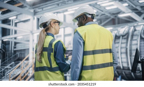 Two Diverse Professional Heavy Industry Engineers Wearing Safety Uniform and Hard Hats Working on Laptop Computer. African American Technician and Female Worker Talking on a Meeting in a Factory. - Shutterstock ID 2035451219