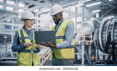 Two Diverse Professional Heavy Industry Engineers Wearing Safety Uniform And Hard Hats Working On Laptop Computer. African American Technician And Female Worker Talking On A Meeting In A Factory.