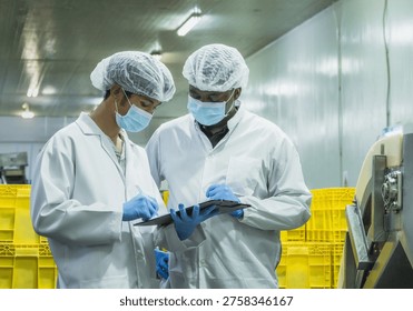 Two diverse quality control inspectors in protective gear discussing production data on a clipboard in a food manufacturing plant to maintain global compliance and operational excellence.