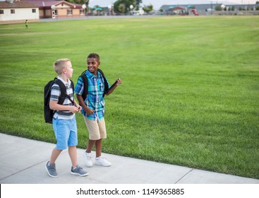 Two Diverse School Kids Walking Home Together After School And Talking Together. Back To School Photo Of Diverse School Children Wearing Backpacks 