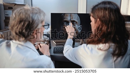 Two diverse scientists discuss 3D model of human head anatomy displayed on PC and tablet computer screens, conduct historical research. Archaeologists or paleontologists work in archaeological lab.
