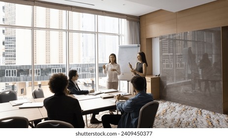 Two diverse speaker women presenting marketing report on whiteboard to coworkers, managers, speaking at flipchart, giving training, presentation in modern office meeting room interior - Powered by Shutterstock