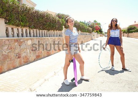 Two diverse teenager friends enjoying a summer holiday together in a suburban street being sporty with skateboard, hoola hoop and headphones technology. Retro adolescent lifestyle outdoors.