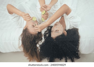 Two diverse woman in bathrobe doing face treatment with facial mask sheet and put cucumber on eyes lying on bed. Beauty and self care concept - Powered by Shutterstock