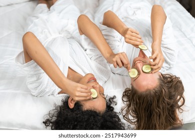 Two diverse women relaxing with cucumber slices on eyes wearing white robes in a spa setting enjoying skincare treatment, self-care routine, beauty and wellness, happy and relaxed, spa day experience - Powered by Shutterstock