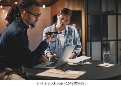 Two Diverse Young Businesspeople Talking On Speakerphone To A Client While Working Together In An Office