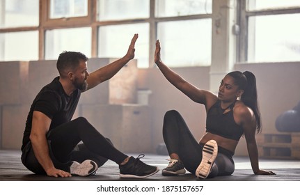 Two diverse young friends in sportswear high-fiving each other while sitting on the floor of a gym