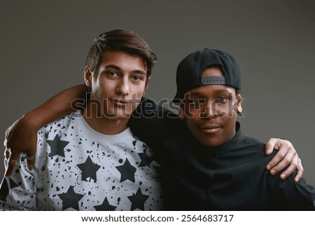 Two diverse young men are posing together with arms around each other, looking at the camera with a cool and friendly attitude in a studio shot against a gray background