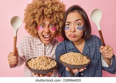 Two Diverse Young Women Pose With Bowls Of Cereals And Spoons Awake Early In Morning Have Outraged Expressions Apply Hydrogel Patches Under Eyes To Reduce Puffiness And Bags Dressed In Nightwear