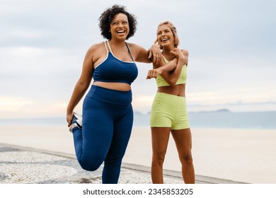 Two diverse young women, wearing sportswear, smile as they stretch and warm up on the promenade. Interracial female friends enjoying their beach workout routine with ocean waves in the background. - Powered by Shutterstock