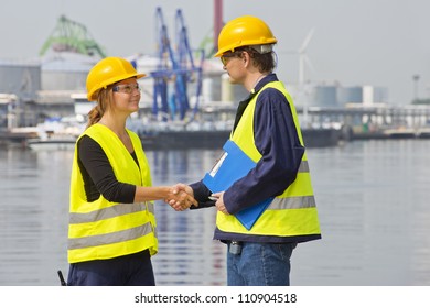 Two Dockers Greeting Eachother In An Industrial Harbor, Wearing The Necessary Safety Gear