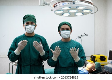 Two Doctors In Operating Room Wearing Green Mask Surgical Gown Surgeon Hair Cap Latex Glove One Stand In Front Of Bed And Electrical Defibrillator Other.