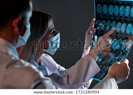 Two doctors in protective masks look and discuss an X-ray or MRI scan of the patient’s brain, isolated on a dark gray background.