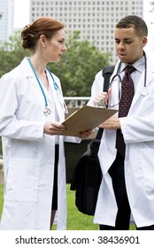 Two Doctors Reviewing A Chart Outside With The Medical Center In The Background.