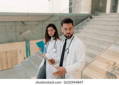 Two doctors with stethoscopes around their necks are walking down stairs in a modern hospital, discussing a patient's chart and medical records