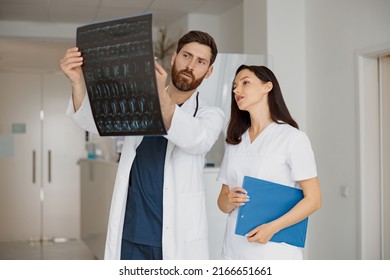 Two Doctors In Uniform Look And Discuss An X Ray Or MRI Scan Of The Patient Spine